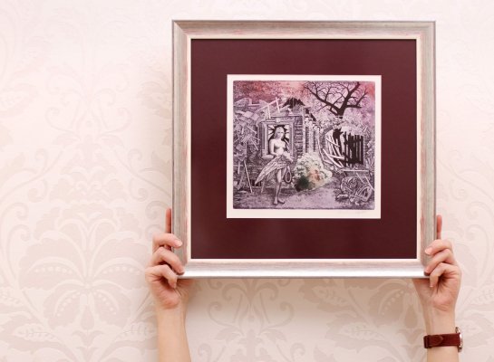 Ruce drží zarámovaný kolorovaný lept Josefa Dudka v německém stříbrném rámu se zlatem a vínovou paspartou v interiéru galerie Frame-it.cz v Praze 7.