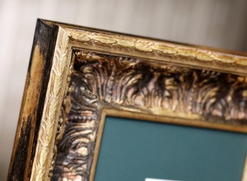 Květinový ornament s akantem a vavřínovými listy na mohutném barokizujícím černo-zlatém rámu s částí tmavozelené pasparty (zarámovaný tisk není vidět).