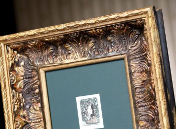 Detailní pohled na horní polovinu zarámovaného leptu Jindřicha Pilečka ve velkorysé tmavozelené paspartě a velmi mohutném černo-zlatém rámu s barokizujícím květinovým ornamentem v luxusním interiéru se zlatými tapetami.