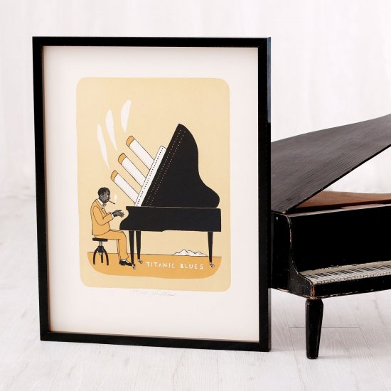 Litografie Jiřího Slívy Titanic Blues zarámovaná v moderním černém lesklém rámu s prostorovým efektem s motivem pianisty je vyfotografovaná vedle miniaturního klavíru.
