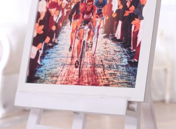 Plakát Paris-Roubaix od Luke Tomski zarámovaný bez paspartování v bílém borovicovém rámečku s viditelnou kresbou letokruhů borovicového dřeva s šedomodrými hranami na stojanu v interiéru – pohled na dolní polovinu plakátu.