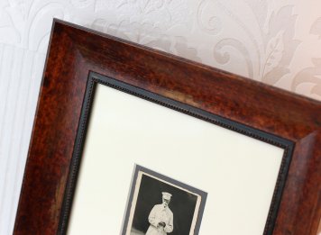 Výsek elegantního historizujícího dřevěného rámu tvaru oblouku s šelakovou patinou, svrchní bílou a spodní tmavošedou paspartou a částí zarámované fotografie prezidenta Masaryka na procházce v Lánech.