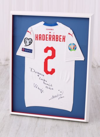 Bílý hluboký rám na reprezentační dres fotbalisty Pavla Kadeřábka na modrém pozadí s červenými vnitřními boky s viditelnými nášivkami UEFA na složených rukávech.