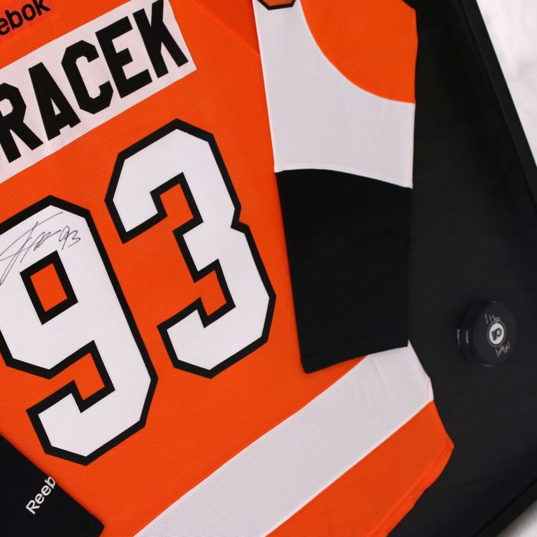 Pohled zblízka na podepsaný hokejový NHL dres Jakuba Voráčka Philadelphia Flyers na černém podkladu a v rámu společně s podepsaným pukem v unikátním muzeálním držáku.