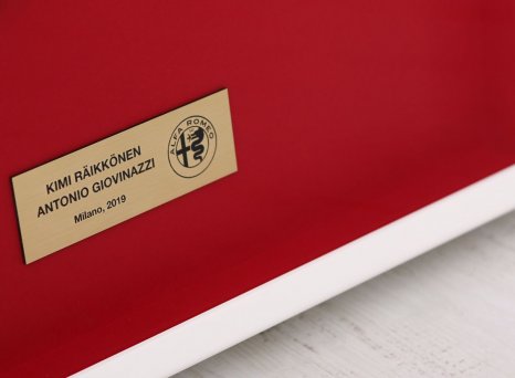 Vysvětlující gravírovaná cedulka s logem Alfa Romeo u rámované závodní kukly Formule 1