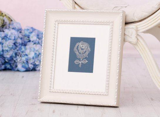 Paličkovaná krajka květiny na modrém podkladu adjustovaná s lomenou bílou paspartou a širším krémovým italským rámem se stříbrnými ornamenty na hranách.