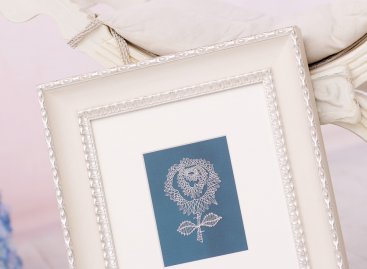 Detailní pohled na jemnou bílou paličkovanou krajku ve tvaru květiny na modrém podkladu, zarámovanou s krémovou paspartou ve zdobeném toskánském rámu. 