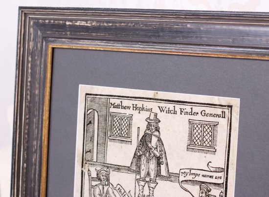 Detail profilovaného černého rámu, který je kartáčovaný až na surové dřevo, voskovaný a patinovaný do starého vzhledu. Rám je doplněný o šedou paspartu. Vidět je část zarámovaného tisku z knihy The Discovery of Witches.