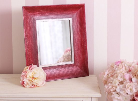 Malé tmavočervené kosmetické zrcadlo na stole v pokoji s tapetami a květinami, široký dřevěný rám s patinou a stříbrnou vnitřní linkou.
